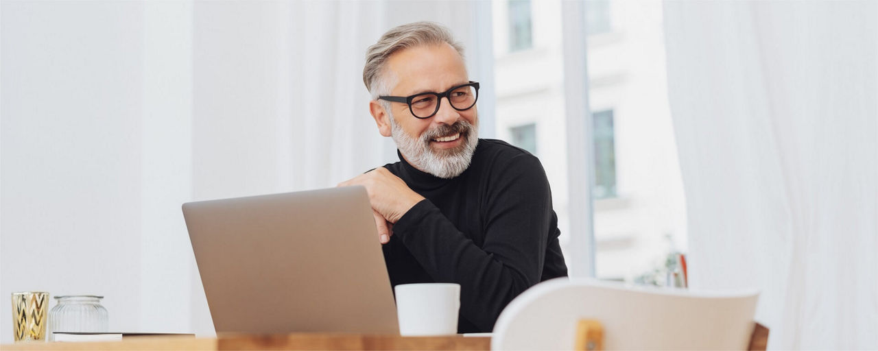 Ein lachender Mann am Schreibtisch mit Laptop und einer Tasse