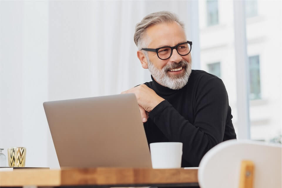 Ein lachender Mann am Schreibtisch mit Laptop und einer Tasse