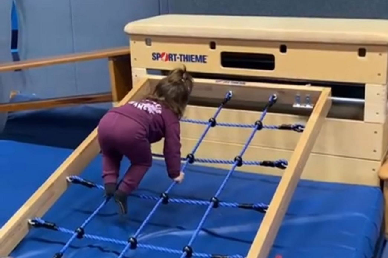 Kleines Kind klettert in der Turnhalle auf einem schrägen Kletternetz, gesichert durch blaue Weichbodenmatten.