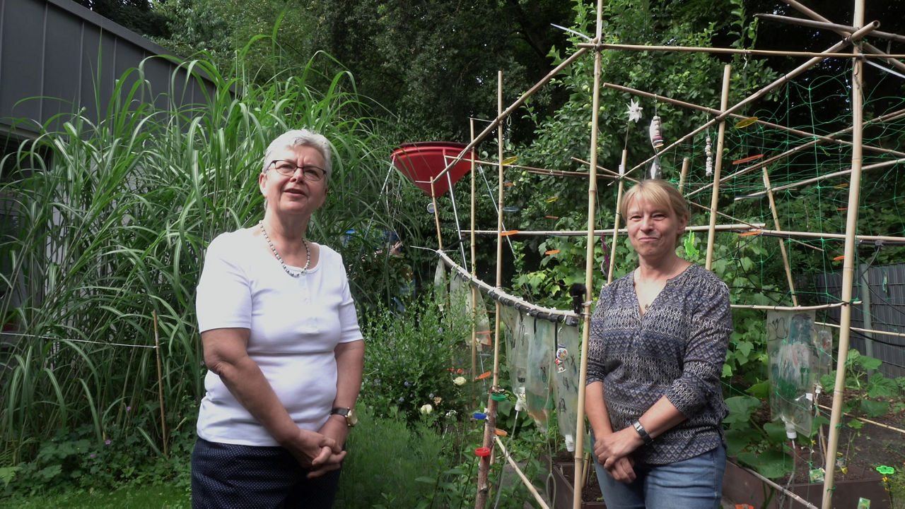 Zwei Frauen von der Kindertagesstätte stehen in einem grünen Nutzgarten und lächeln freundlich in die Kamera. Sie vertreten die Kindertagesstätte Lothringer Straße und haben beim Klimahelden 2024 im Bereich Vereine den 2. Platz belegt.