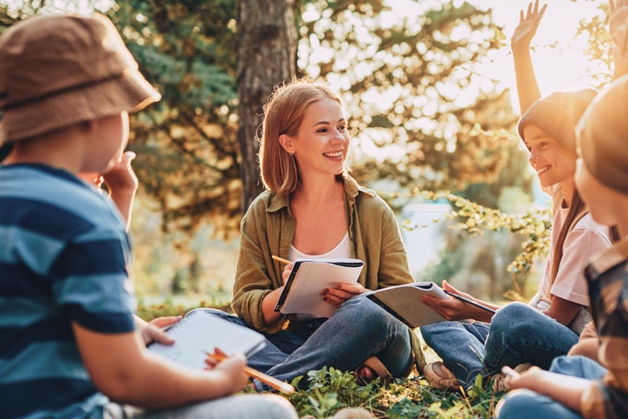 Eine junge Frau und mehrere Kinder sitzen in einem Wald auf dem Boden im Kreis. Alle halten ein Notizbuch in der Hand. Sie redel und lächeln.