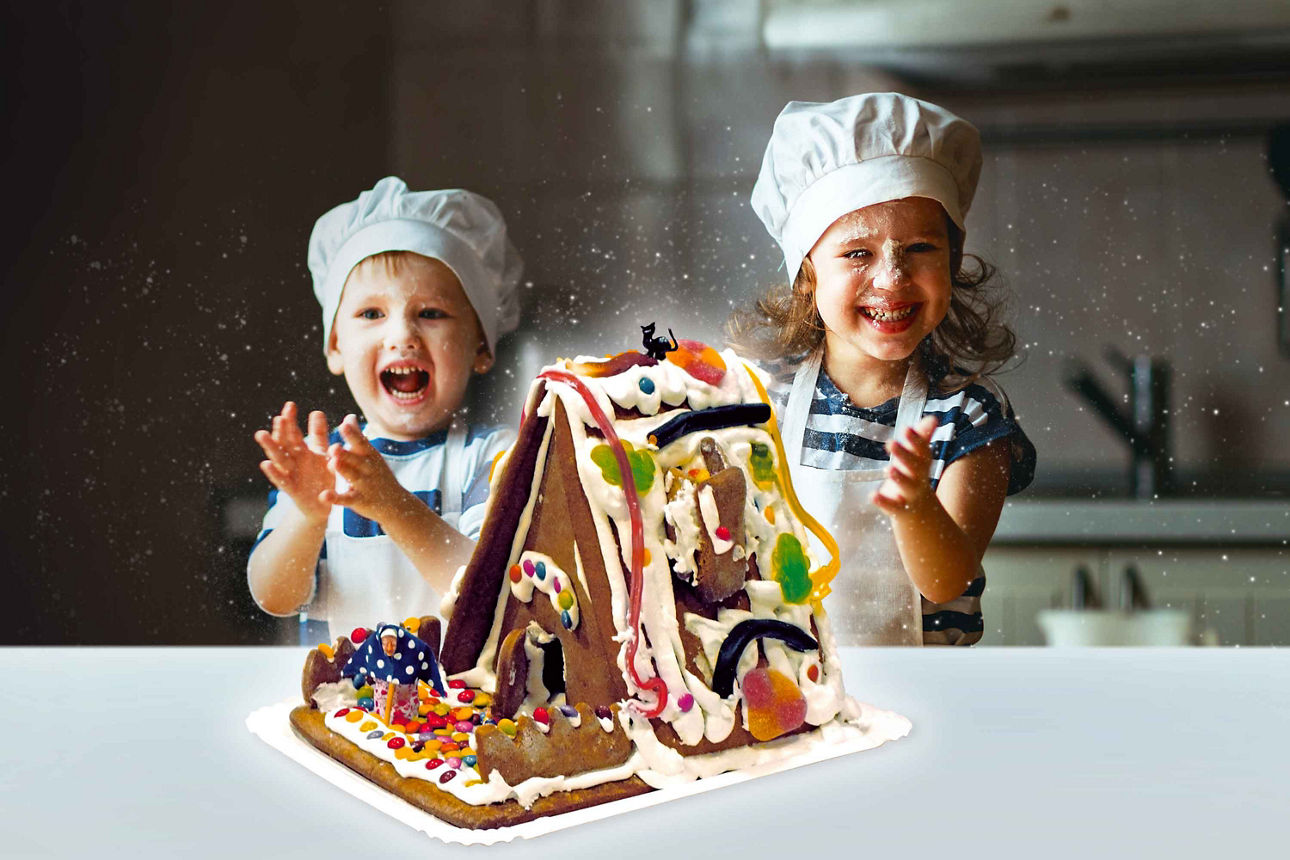 Zwei Kinder mit Kochmützen freuen sich über ein bunt verziertes Lebkuchenhaus in einer Küche.