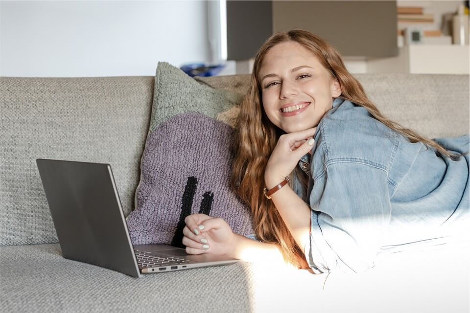 Junge Frau liegt auf dem Sofa auf dem Bauch und ein Laptop steht vor ihr