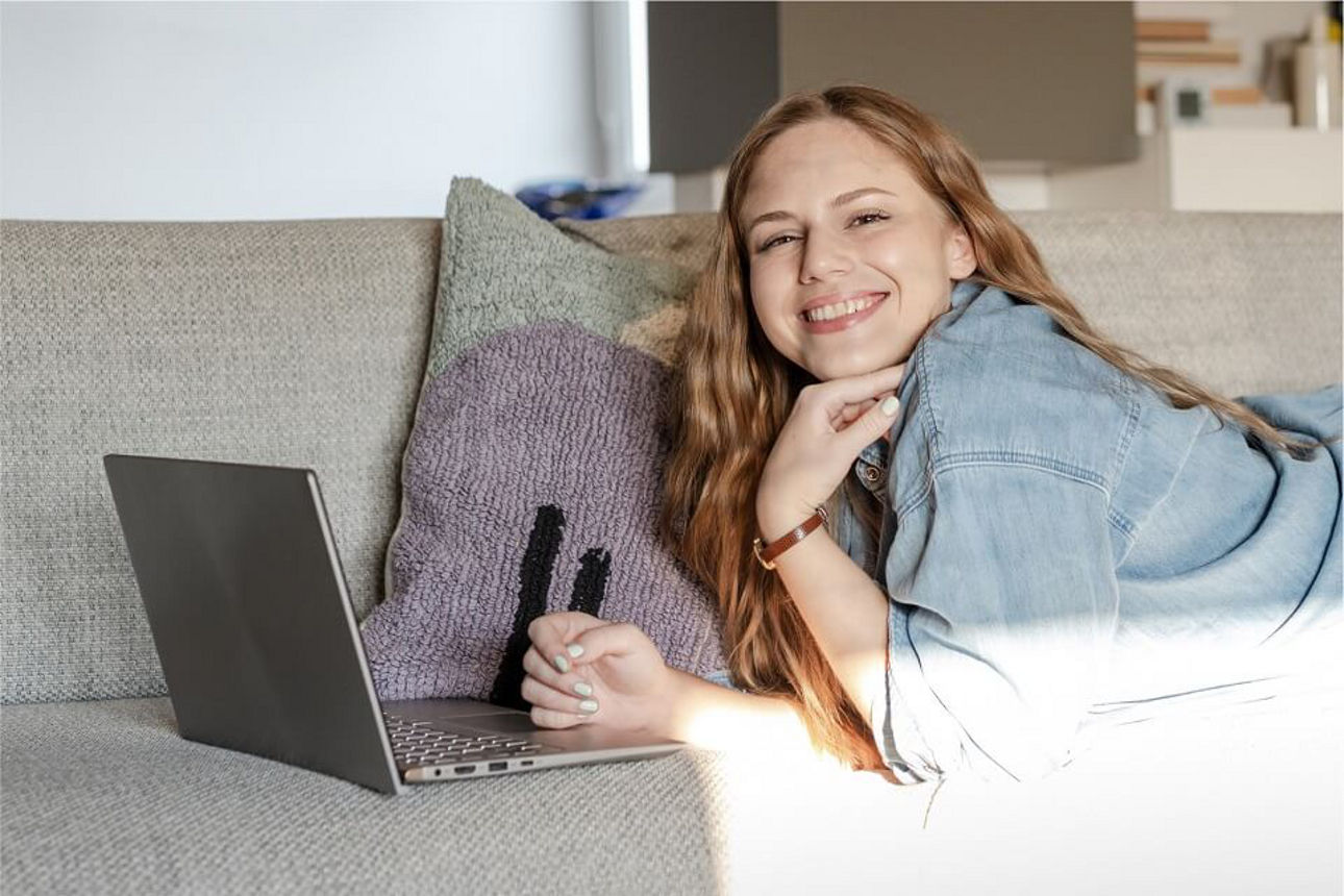 Junge Frau liegt auf dem Sofa auf dem Bauch und ein Laptop steht vor ihr