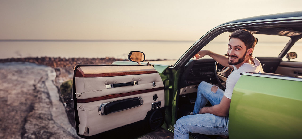 Junger Mann sitzt in einem grünen Auto am Meer mit Sonnenuntergang und lächelt in die Kamera