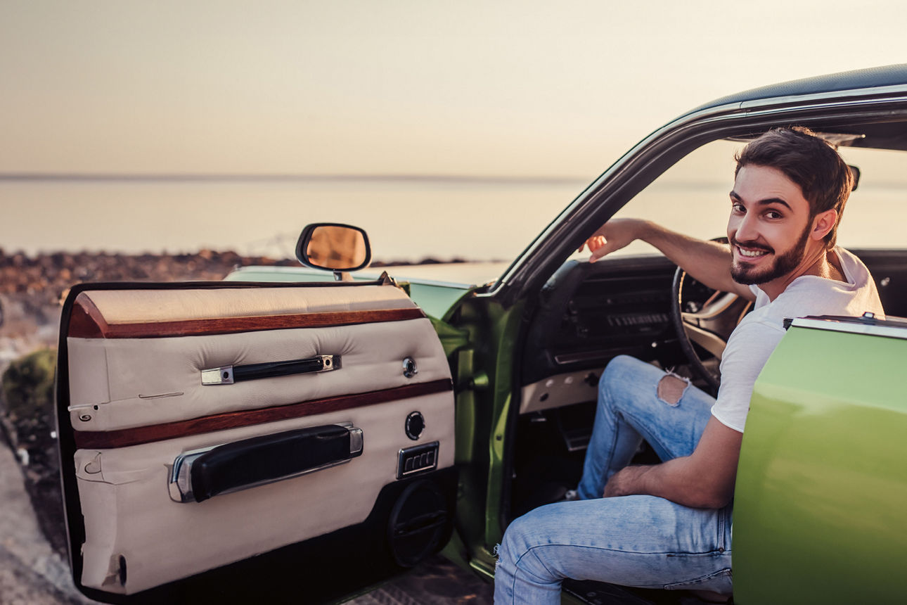Junger Mann sitzt in einem grünen Auto am Meer mit Sonnenuntergang und lächelt in die Kamera