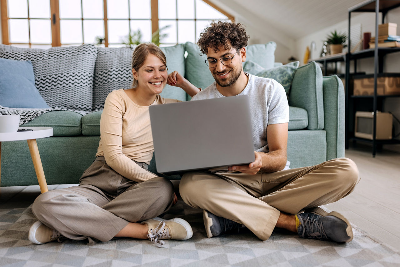 Junges Paar sitzt zu Hause vor dem Sofa und schaut auf einen Laptop