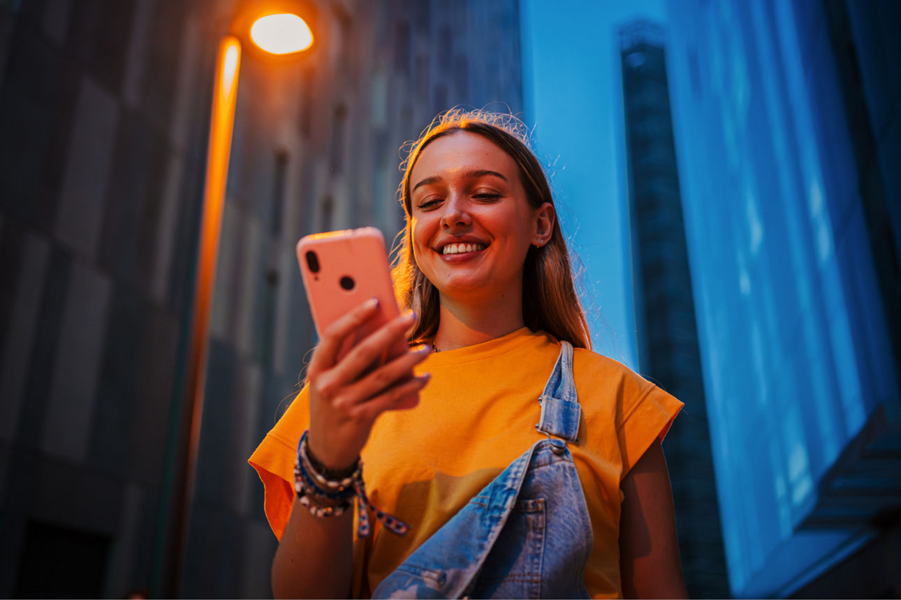 Junge Frau steht zwischen Hochhäusern und schaut lächelnd auf ihr smartphone