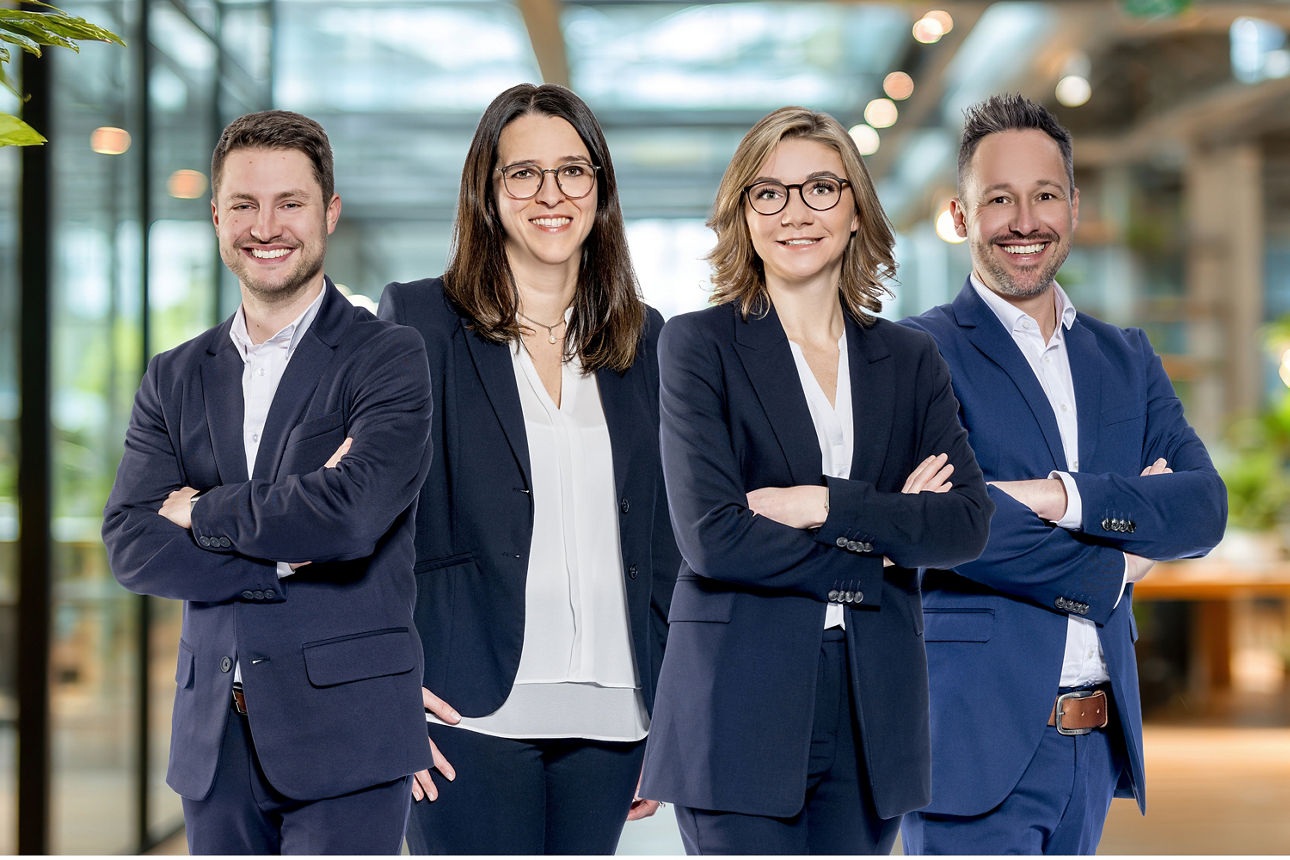 Vier Personen in Businesskleidung stehen mit verschränkten Armen in einem modernen Bürogebäude