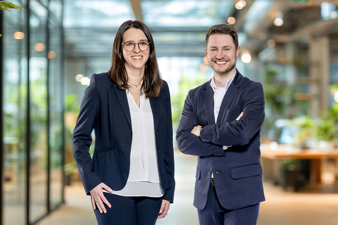 Zwei Personen in Businesskleidung stehen nebeneinander in einem modernen Büro mit großen Fenstern und Pflanzen im Hintergrund