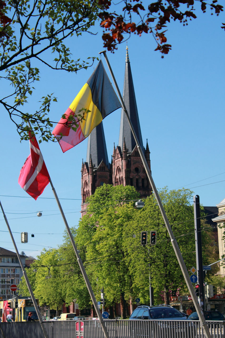 Zwei Flaggen wehen im Wind. Im Hintergrund sind grüne Bäume und zwei hohe spitze Gebäude zu sehen