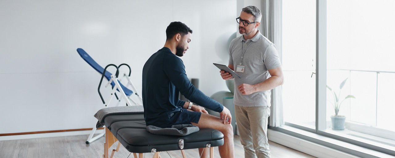 Mann sitzt auf liege und beschreibt Therapeuten Probleme am Knie.