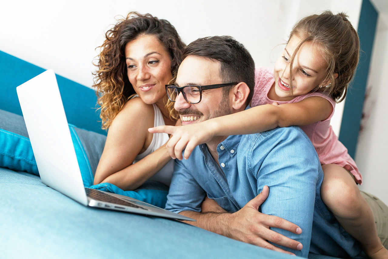 Mutter und Vater liegen mit Tochter auf dem Sofa und schauen sich etwas auf dem Laptop an