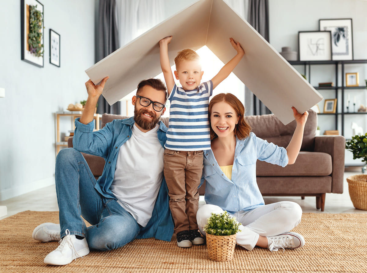 Eine junge Familie sitzt in ihrem Wohnzimmer und hält sich ein Dach über den Kopf