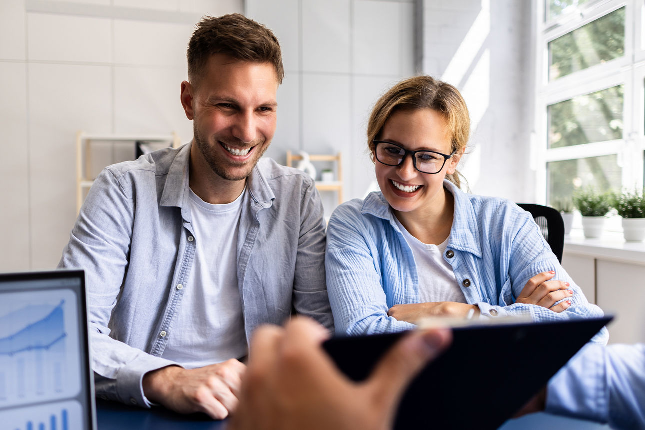Junges Ehepaar sitzen in einem Beratungszimmer und schauen glücklich auf ein Tablet, welches von einem Bankberater gehalten wird