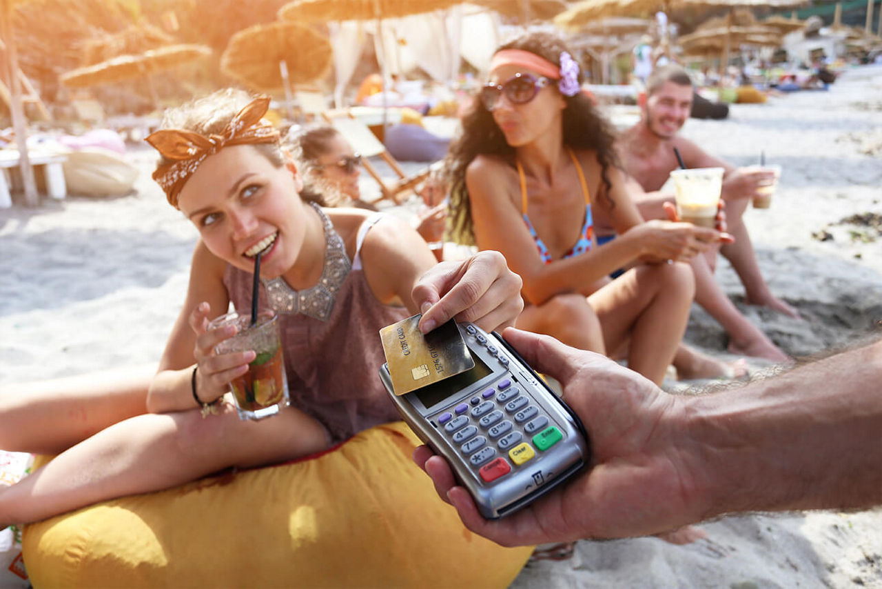 Frauen am Strand und ein Kartenterminal, das von einer Hand ins Bild gehalten wird - eine der Frauen bezahlt mit ihrer Karte