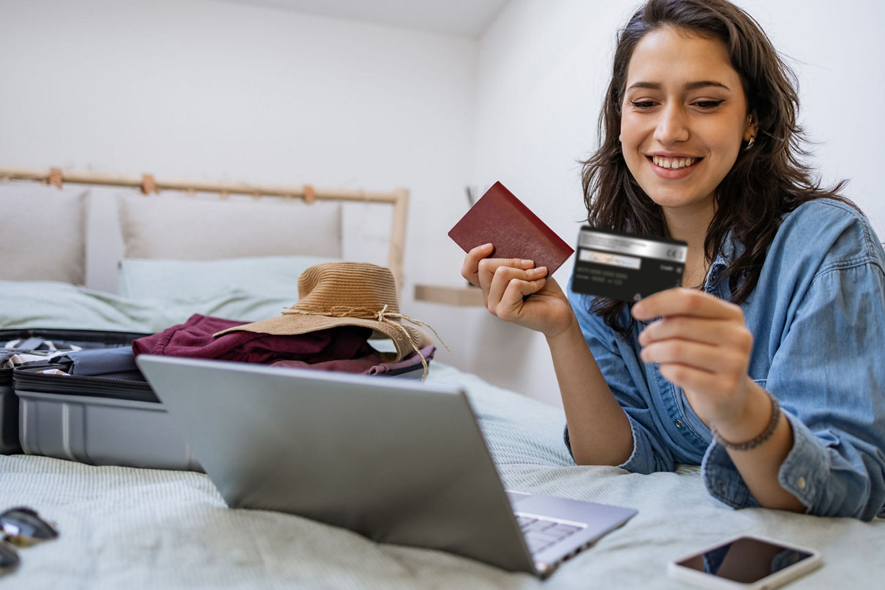 junge Frau hält Reisepass und Kreditkarte in den Händen und schaut auf Laptop, ein gepackter Koffer liegt im Hintergrund