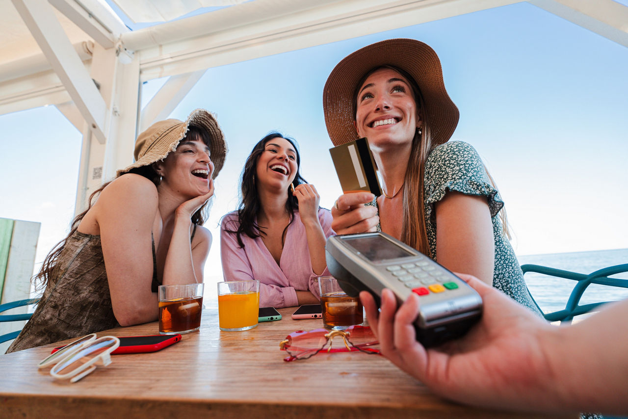 Kreditkarten BesteDrei junge Frauen sitzen in einer Beachbar und bezahlen ihre Drinks mit Kreditkartellstrecke