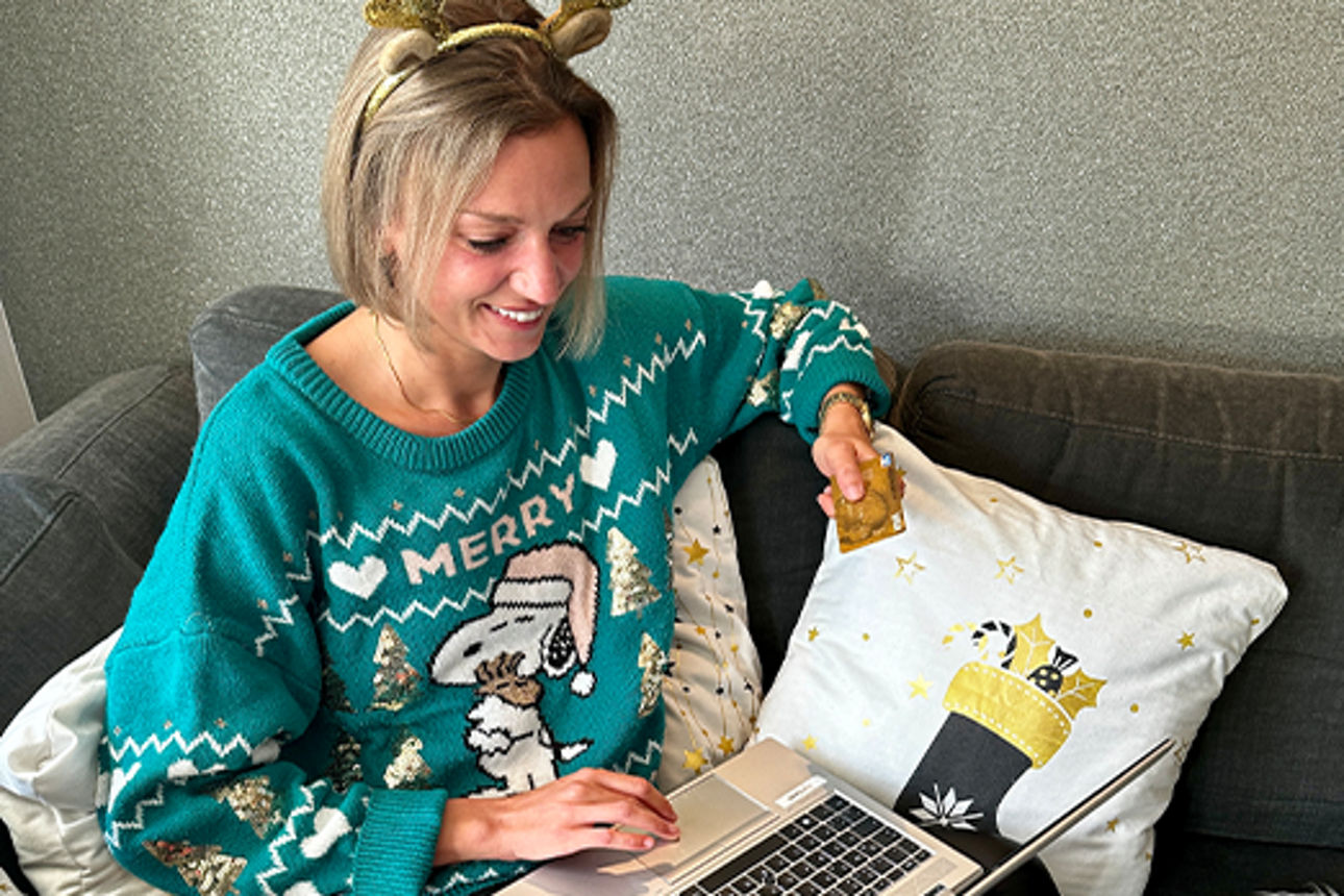 Eine Frau im Weihnachtspulli sitzt auf dem Sofa mit einem Laptop und einer goldenen Kreditkarte in der Hand
