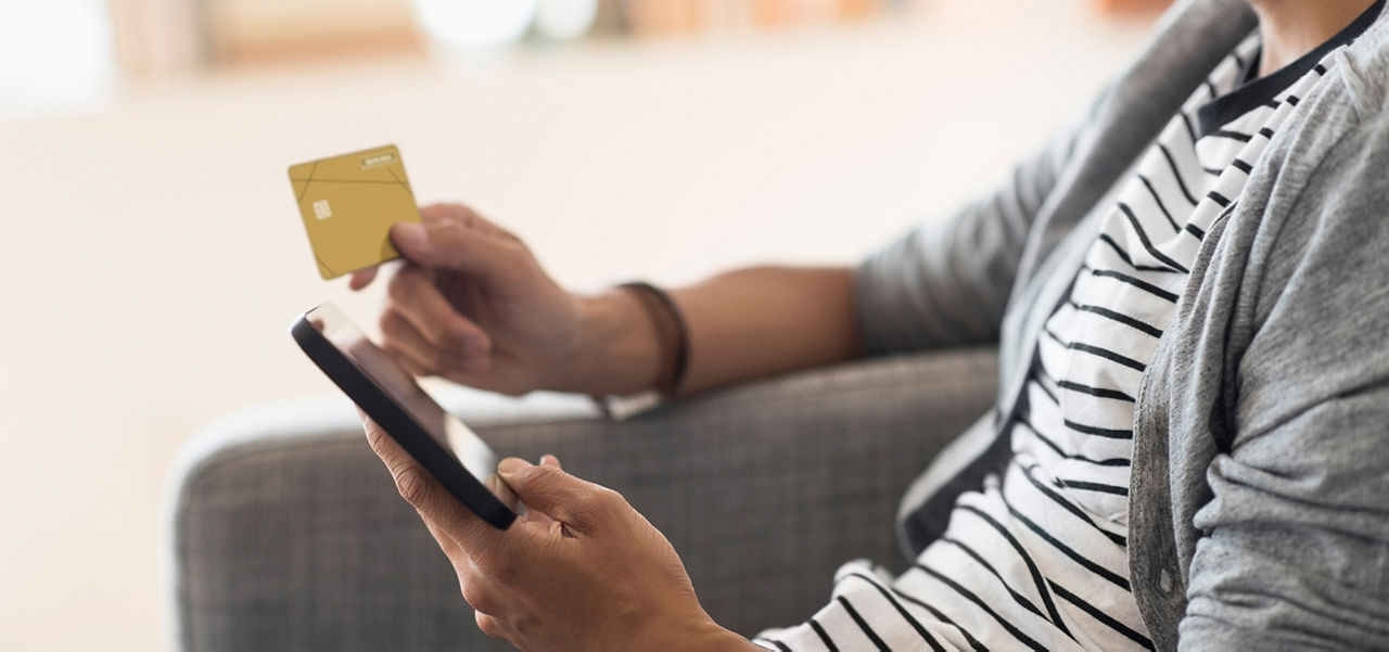 Person sitzt mit einem Smartphone und einer goldenen Kreditkarte in einer Hand auf einem grauen Sofa