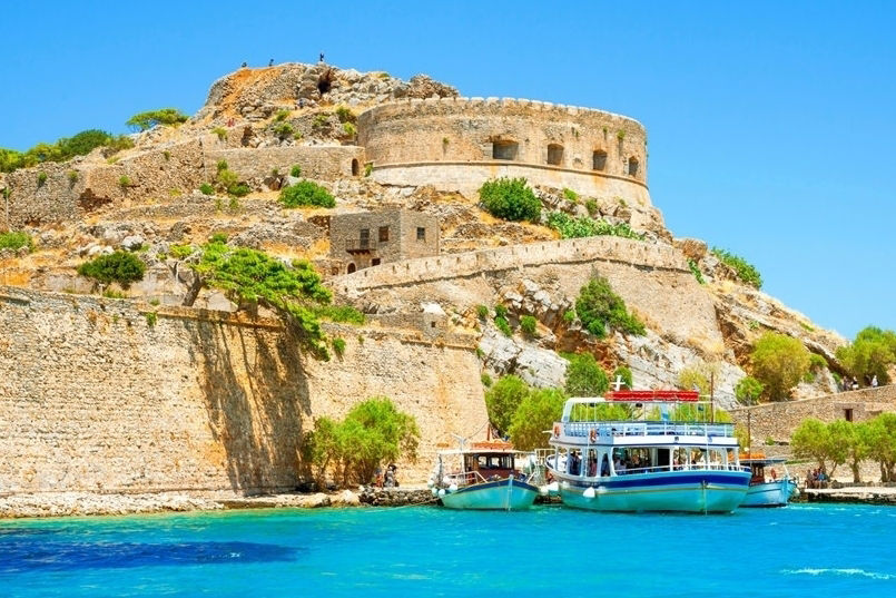 Blick vom Meer auf die Insel Spinalonga, Griechenland mit einer Burg, strahlend blaues Meer und Himme, ein Boot steht am Ufer