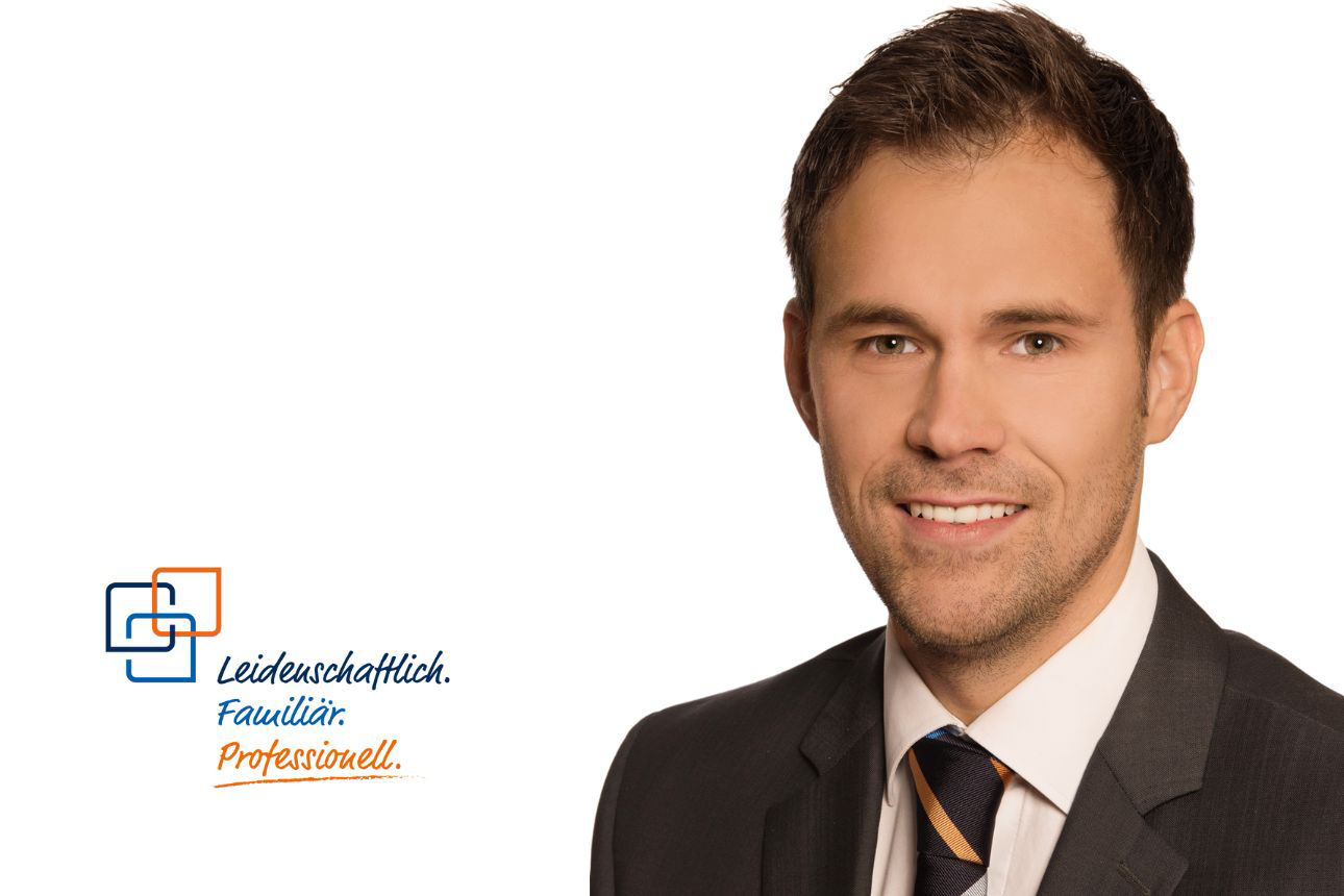 Johann Kroll, lächelnd, mit kurzen dunklen Haaren und einer Krawatte in Dunkelblau und Orange gestreift.