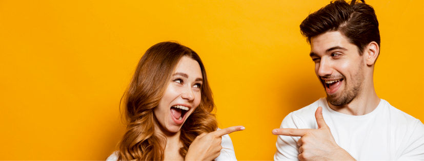 Eine Frau und ein Mann stehen vor einen gelben Wand und zeigen jeweils mit dem Zeigefinger auf sich