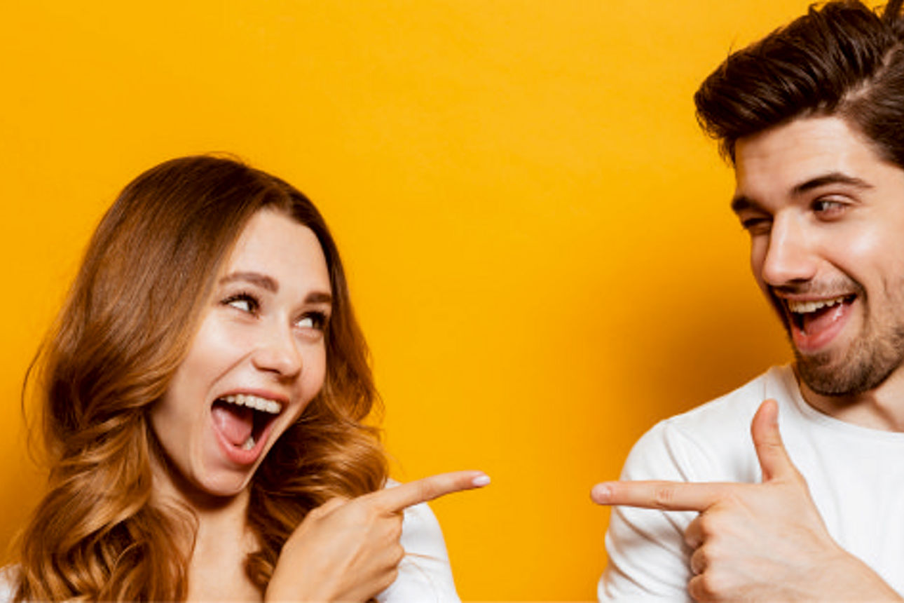 Eine Frau und ein Mann stehen vor einen gelben Wand und zeigen jeweils mit dem Zeigefinger auf sich