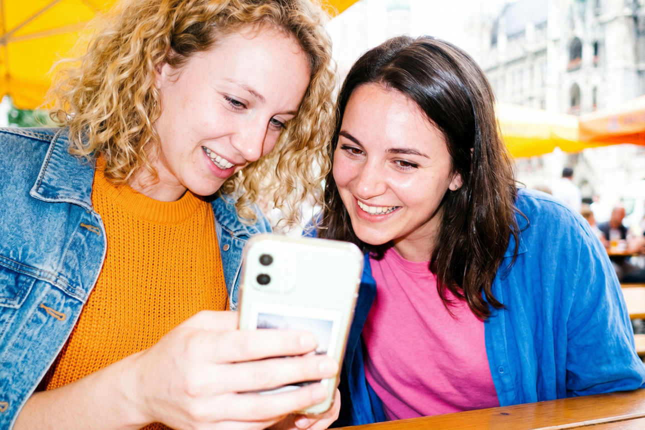 „Zwei junge Frauen sitzen lächelnd an einem Tisch im Freien und schauen gemeinsam auf ein Smartphone.