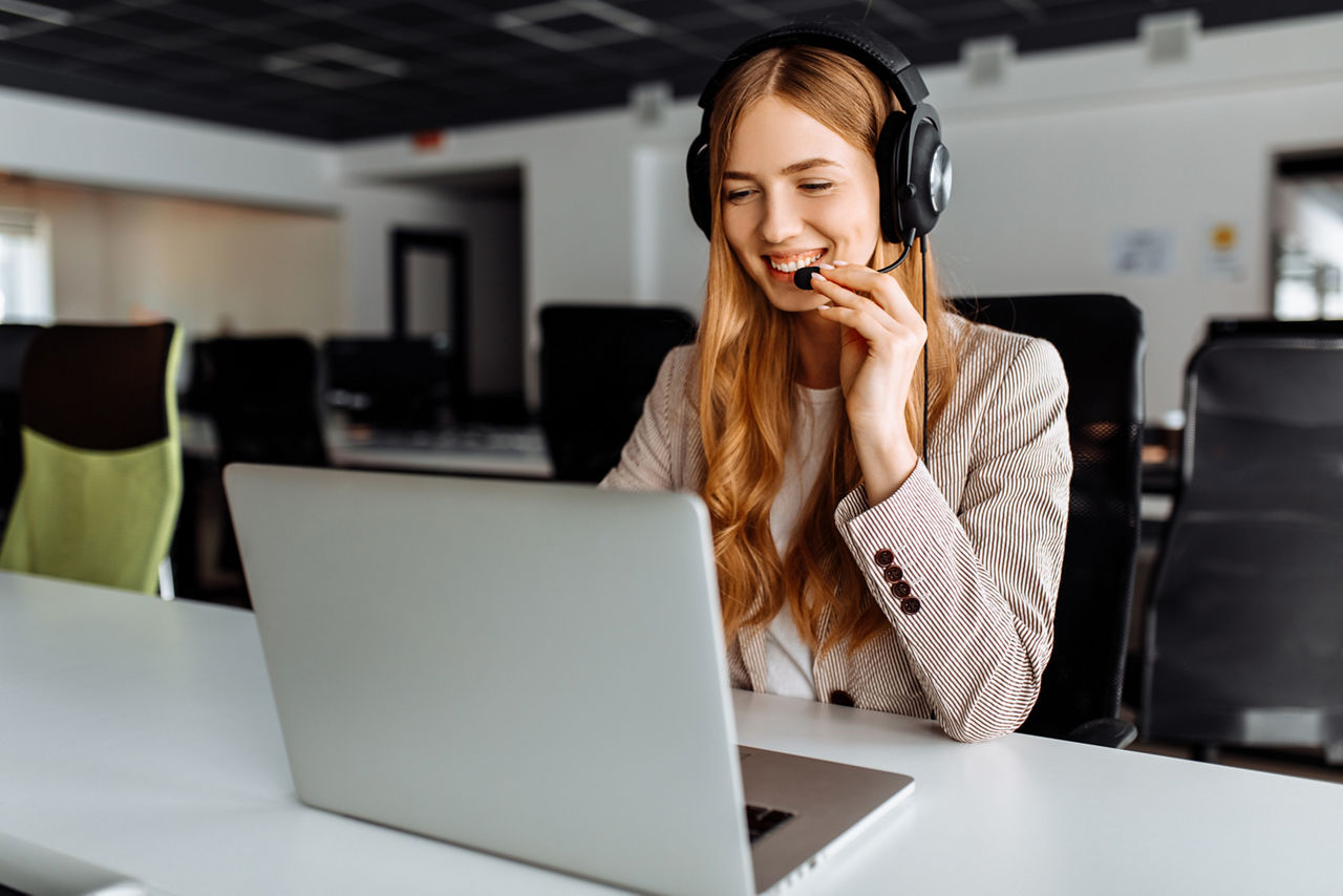 young female operator in headphones with headset working with laptop sitting at table at workplace, technical support concept