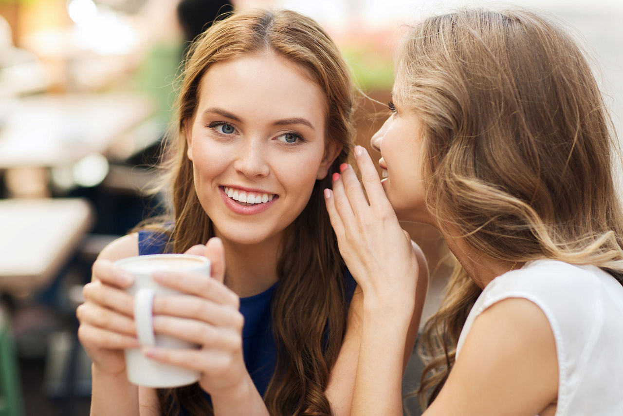 Eine Frau flüstert einer anderen Frau die eine Tasse Kaffee mit beiden Händen hält etwas ins Ohr