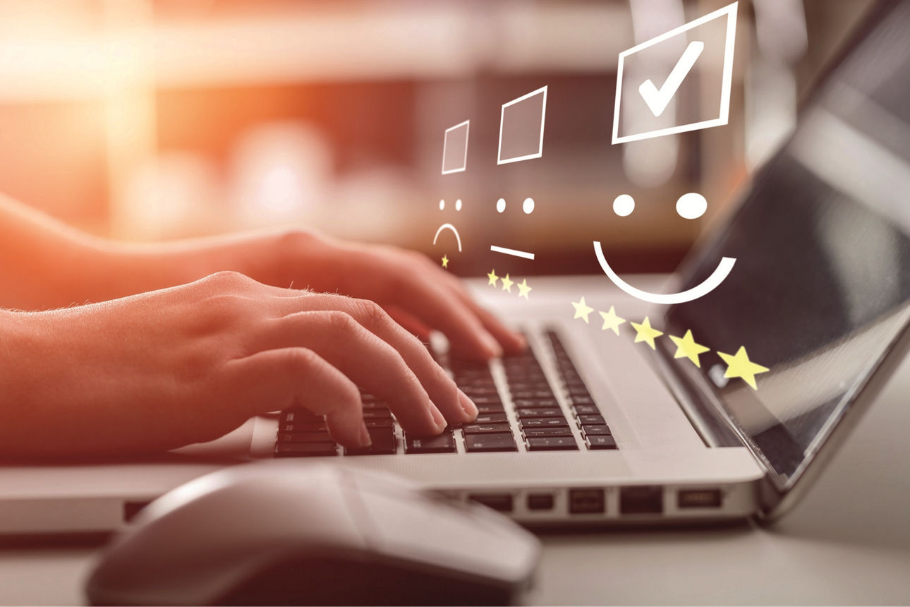 Hände tippen auf einer Laptoptastatur, zu sehen sind eine Maus und verschiedene Smileys mit Sternen für die Kundenumfrage