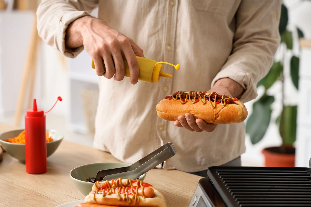 Person hält Hotdog in der linken Hand und gibt mit der rechten Hand Senf aus einer gelben Flasche darauf