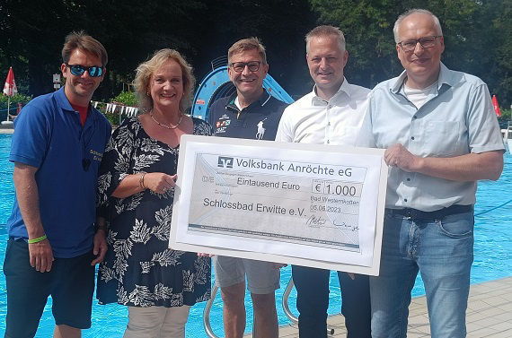 Fünf Personen halten gemeinsam einen großen Scheck neben einem öffentlichen Schwimmbad