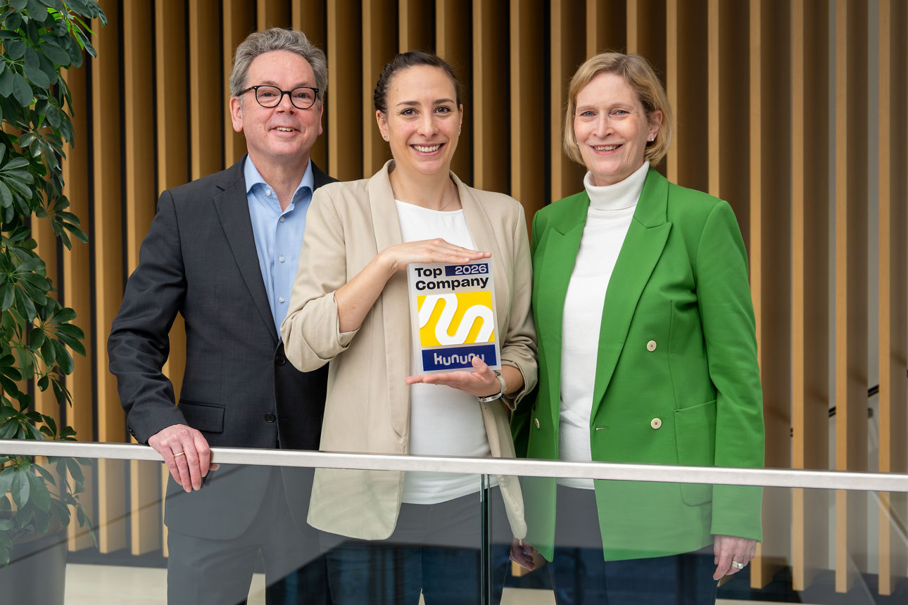 Drei Personen stehen nebeneinander und präsentieren ein „Top Company 2026“-Siegel vor moderner Innenarchitektur.