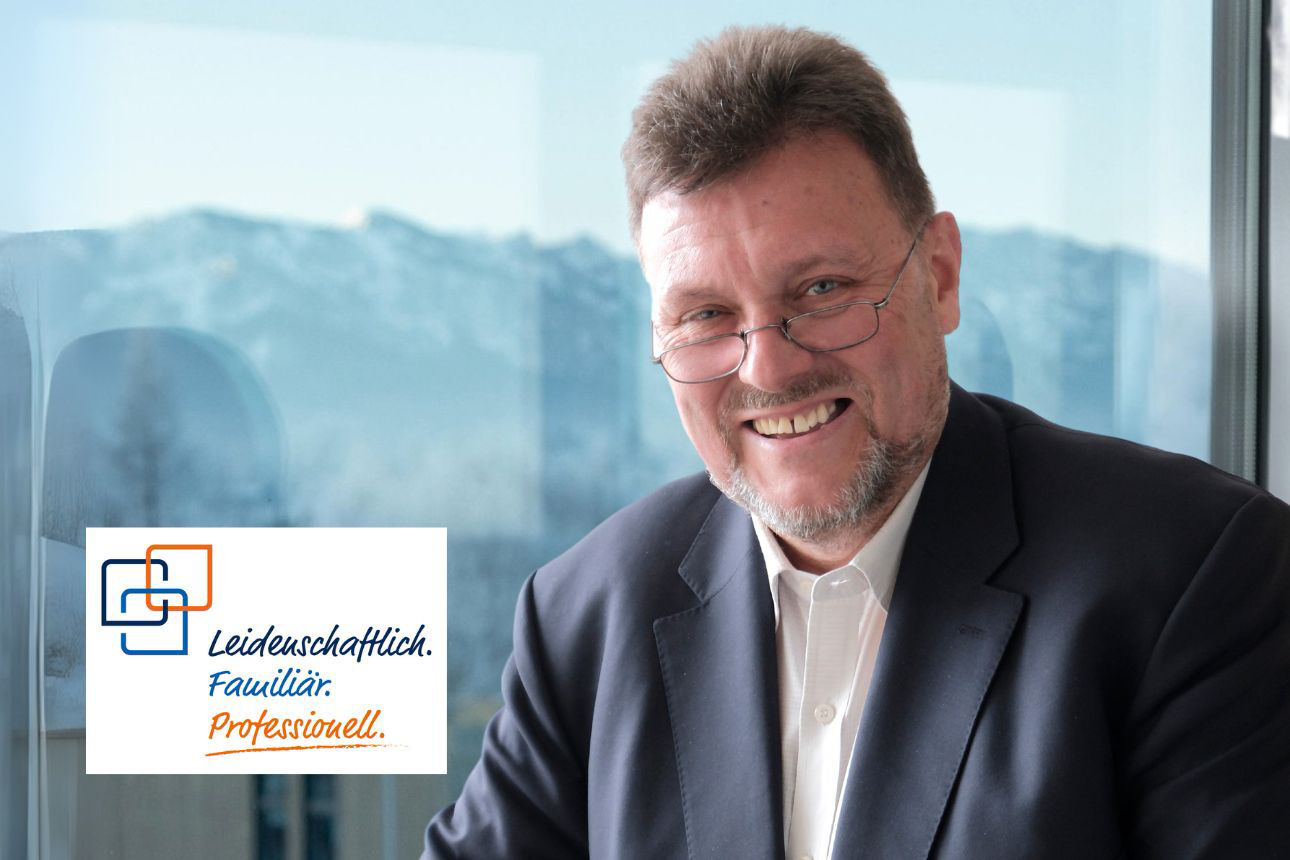 Porträt von Kurt Dirschl, einem lächelnden Mann mit Brille und Bart im dunklen Anzug vor einem Fenster mit Bergpanorama. Daneben das Firmenlogo und der Slogan "Leidenschaftlich. Familiär. Professionell."