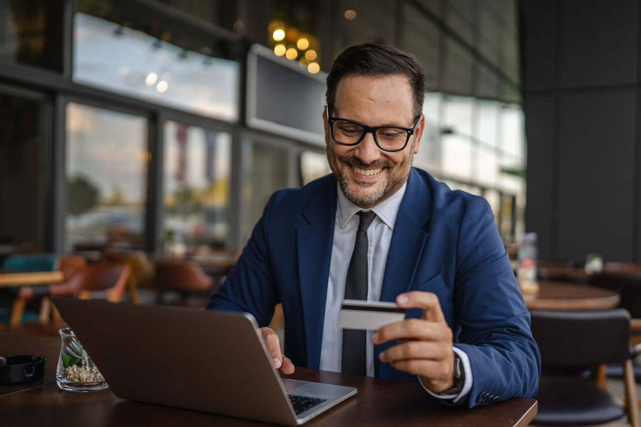 Lächelnder Geschäftsmann sitzt im Cafè vor einem Notebook und hält eine Kreditkarte in der Hand