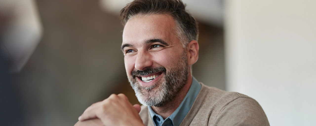 Eine lächelnder Mann sitzt in einem Bürobereich und lehnt einen Arm auf den Tisch. 