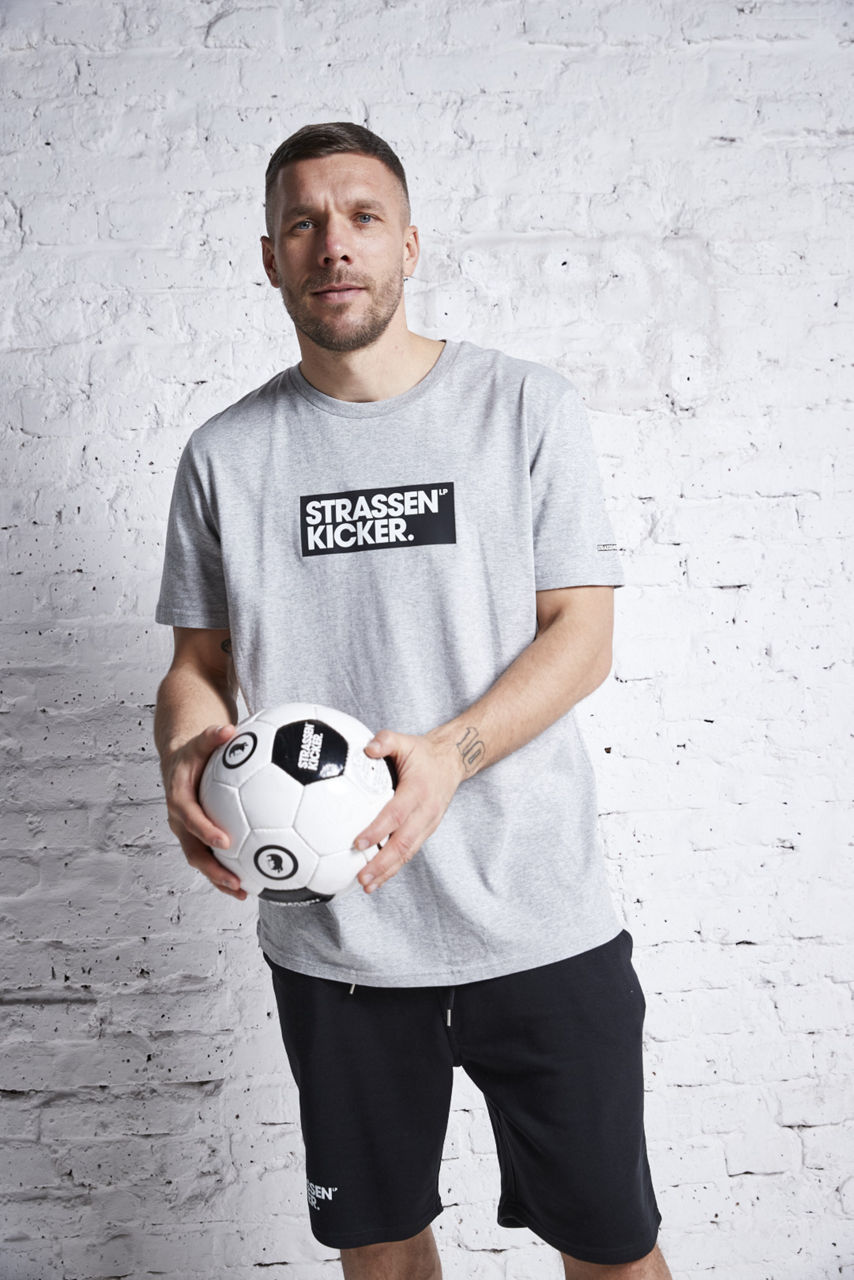 Lukas Podolski hat einen Fuball in der Hand und steht vor weißer Mauer