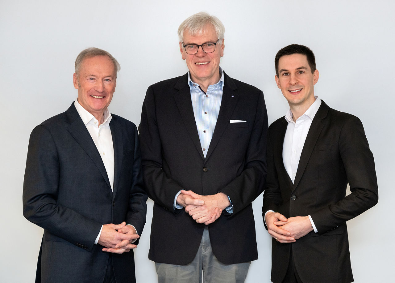 Drei Männer in dunklen Anzügen stehen zusammen vor einem schlichten weißen Hintergrund und strahlen Professionalität und Sel