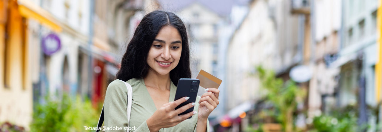 Einen Frau schaut lächelnd auf ihr Handy. In der anderen Hand hält sie eine Kreditkarte 