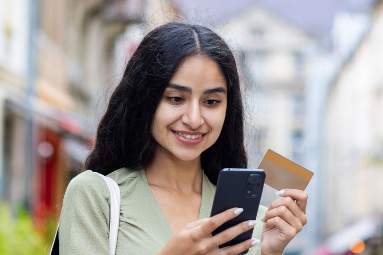 Einen Frau schaut lächelnd auf ihr Handy. In der anderen Hand hält sie eine Kreditkarte 