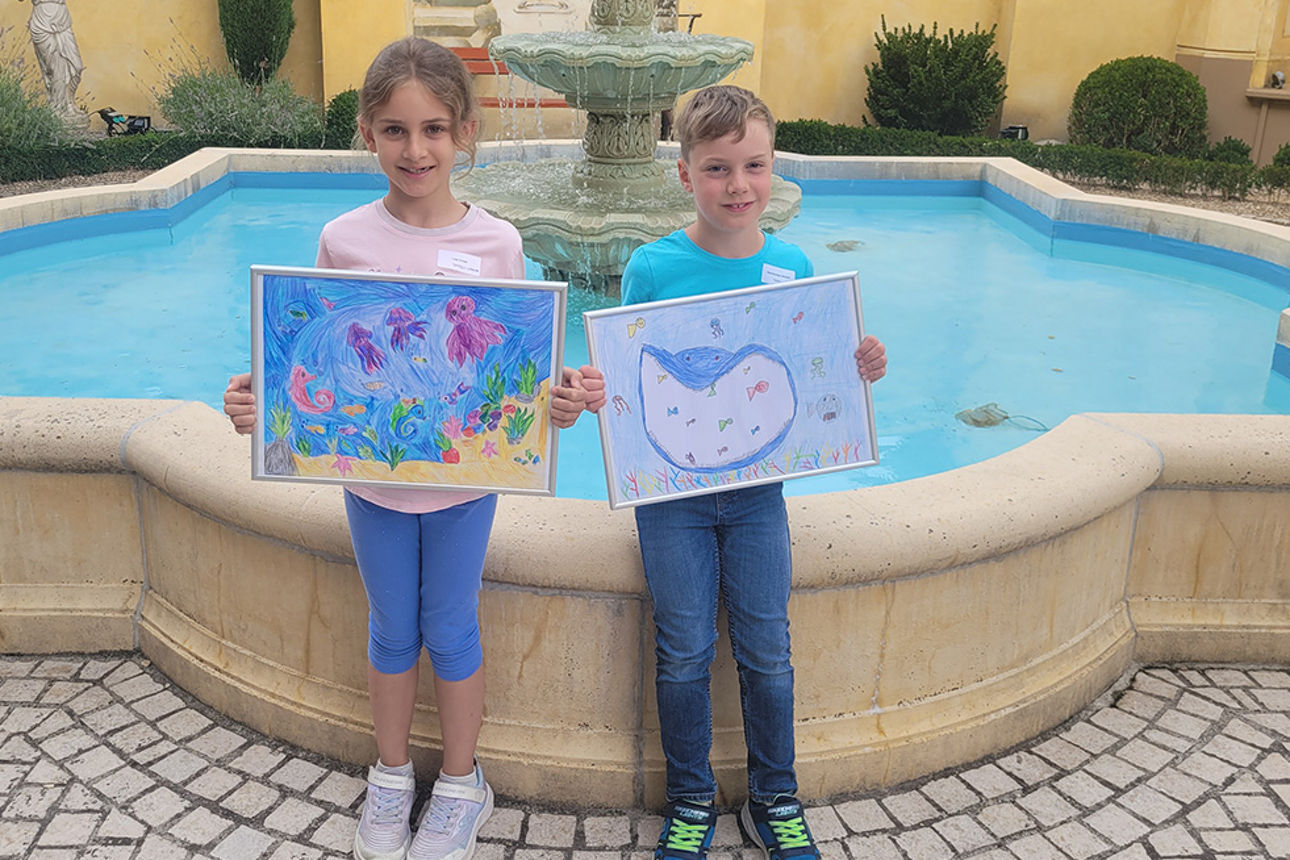 Die beiden Preisträger Luisa Schaaf und Raul Hirschlein Wohlfart stehen mit ihren Bildern vor einem Brunnen
