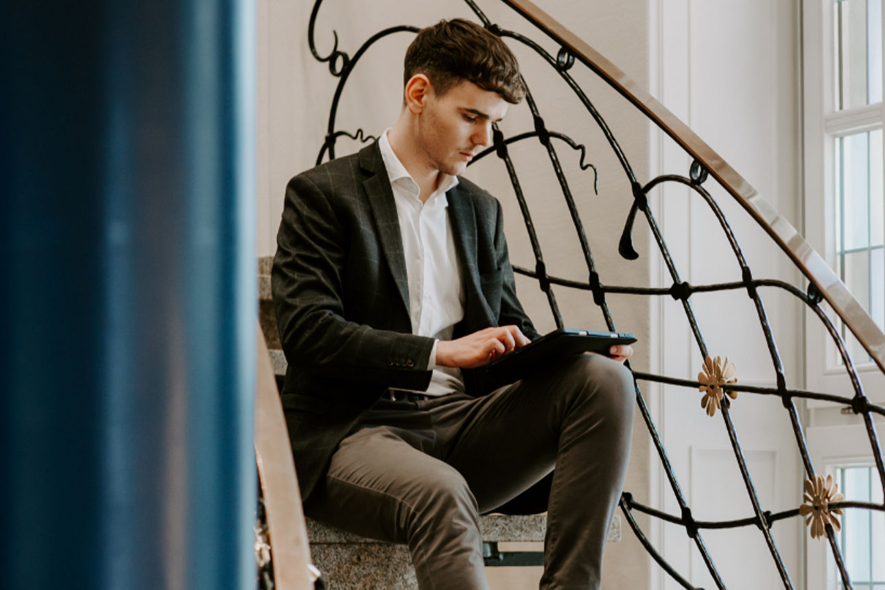 Ein junger Mann sitzt auf einer Treppe und arbeitet konzentriert an einem Tablet.