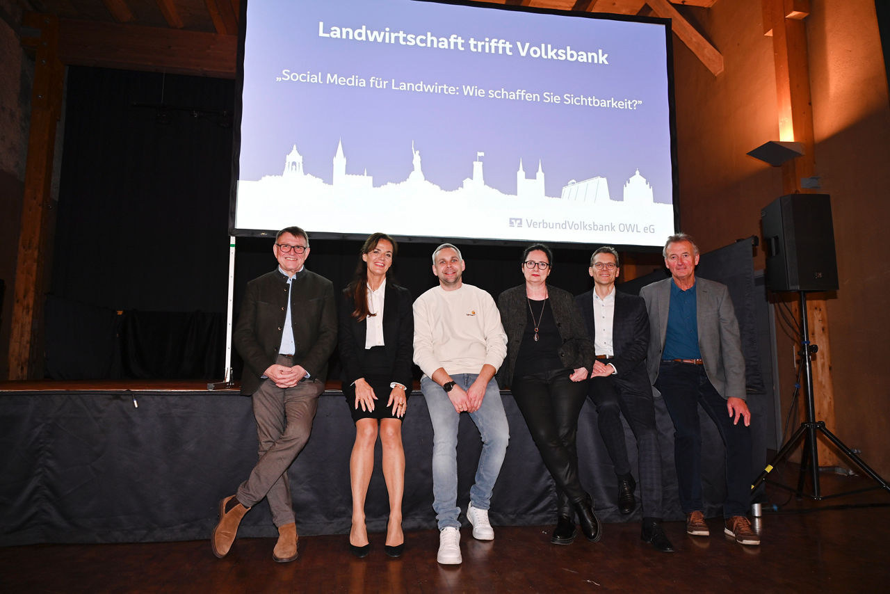Die Beteiligten der Veranstaltung Veranstaltung „Landwirtschaft trifft Volksbank“ im KuStall in Höxter-Ottbergen lehnen vor einer Bühne. 