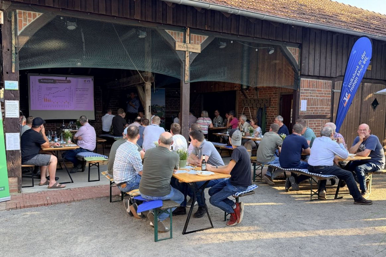 Besucher sitzen an Bierbänken in einem offenen Hof vor einer Scheune und verfolgen eine Präsentation auf einer Leinwand mit D