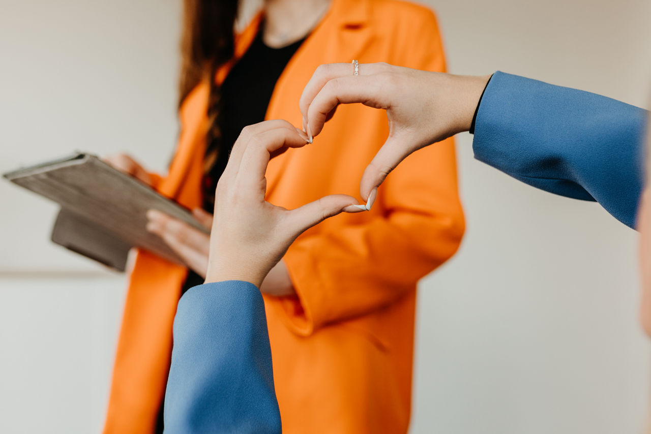 Eine Person zeigt mir ihren Händen eine Herzform und im Hintergrund ist eine Person mit einem orangenen Blazer zu sehen