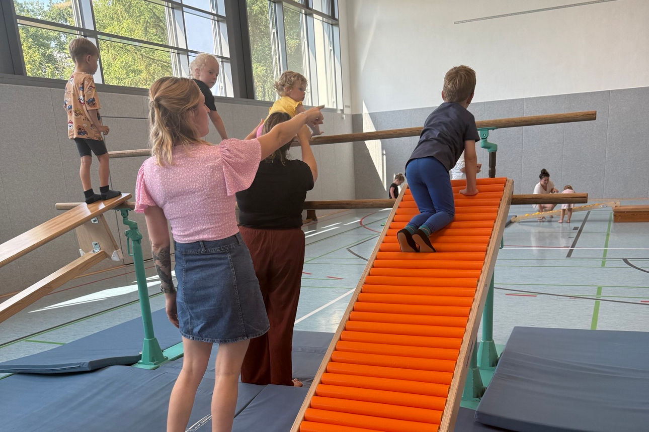Kind klettert in der Turnhalle eine Rollbrettbahn hoch, begleitet von zwei Betreuungspersonen auf Weichbodenmatten.