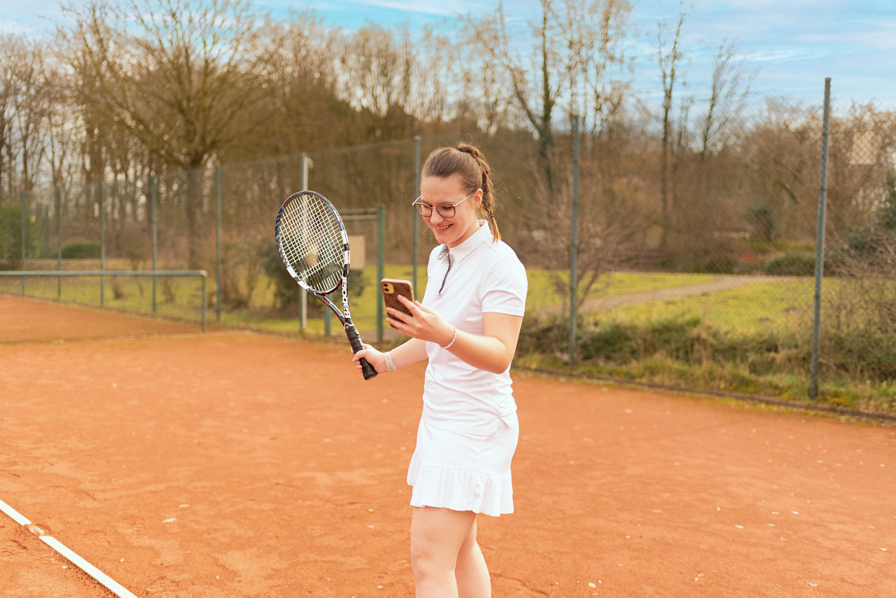 Junge Frau steht mit einem Handy und einem Tennisschläger in der Hand auf dem Tennisplatz
