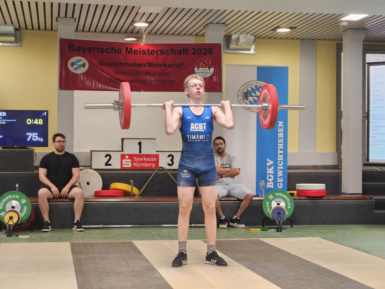 Gewichtheber hebt eine Langhantel mit roten und grünen Gewichtsscheiben in einer Sporthalle während eines Wettkampfs.